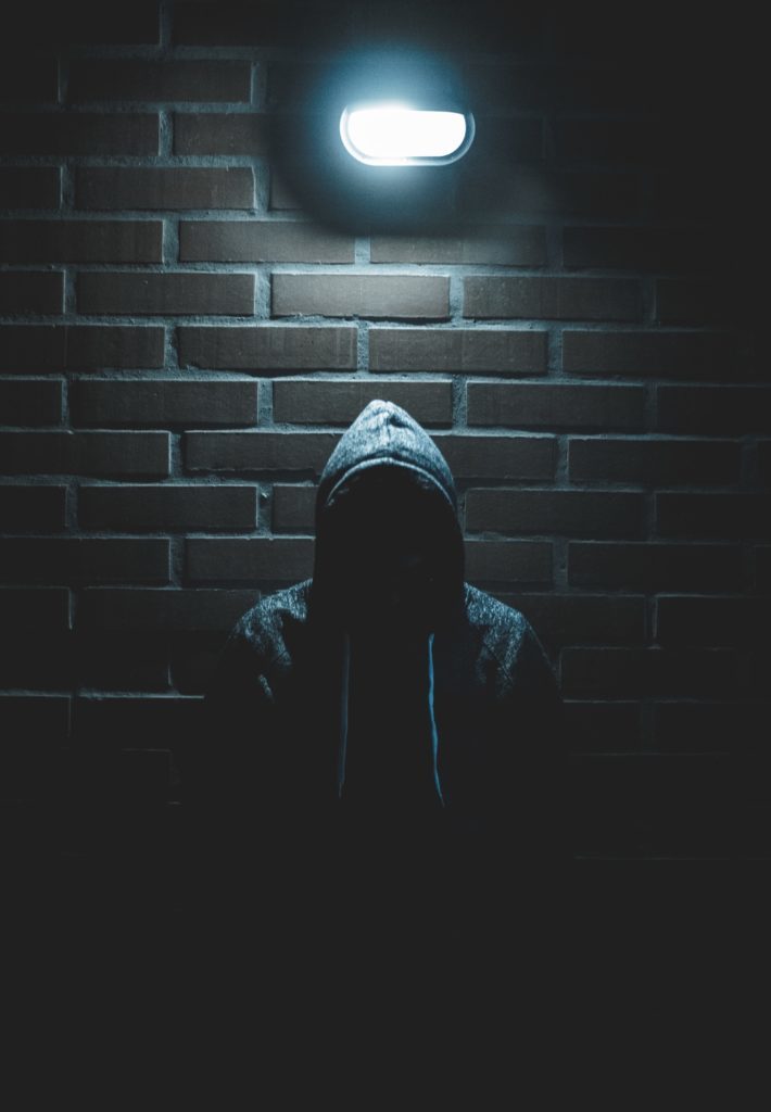 Man in a dark hood against a brick wall with a light above him